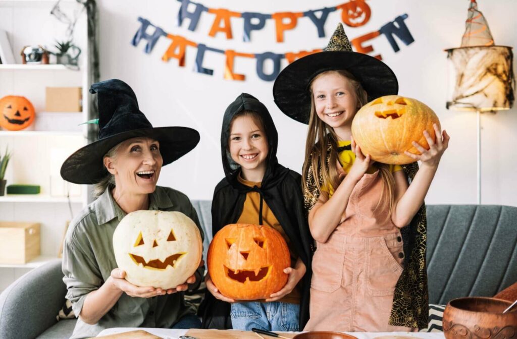 A happy senior dressed up and carving jack-o-lanterns with their 2 grandchildren.