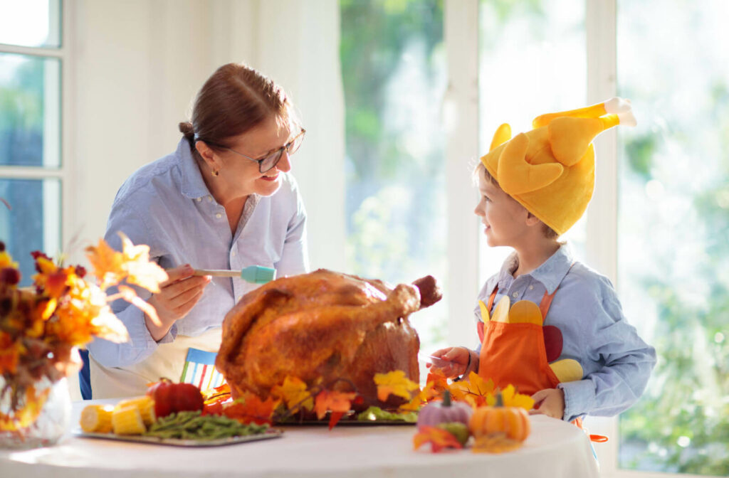 A child wearing a silly turkey-shaped hat laughing with a grandparent while they prepare a Thanksgiving turkey together.